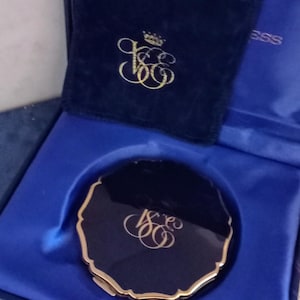 May include: A navy blue compact mirror with a gold-colored scalloped edge and a gold monogram, presented in a matching box with a velvet pouch. The box is lined with blue satin and has the word "ESS" printed on the inside.
