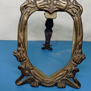 May include: An antique brass-colored picture frame with an oval opening. The frame features ornate floral and scrollwork designs. It has a small stand on the back to allow it to stand upright. The frame is approximately 5 inches tall.