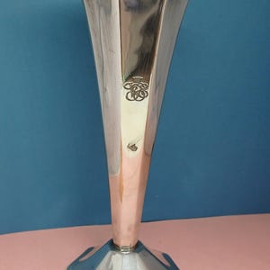 Peut inclure: Un vase argenté avec un dessus évasé et une base hexagonale. Le vase a un corps mince et allongé et un emblème décoratif près du sommet. Le vase est présenté sur un fond bleu et rose.