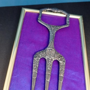 Könnte beinhalten: Eine verzierte, silberfarbene Serviergabel mit drei Zinken und einem Griff, präsentiert auf einem violetten Samthintergrund in einem goldgerahmten Rechteck. Die Gabel weist detaillierte Blumen- und Scrollwork-Designs auf.