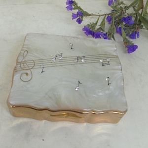 May include: A rectangular, off-white trinket box with a gold trim. The lid features a musical staff with notes and sparkling accents. A gold treble clef is on the left side. A sprig of purple flowers rests nearby.