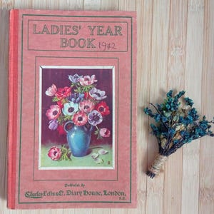 May include: A vintage 1942 Ladies' Year Book with a dusty rose cover, featuring a central illustration of a blue vase filled with colourful anemone flowers. The text reads Ladies' Year Book 1942 and Published by Charles Letts & Co, Diary House, London.
