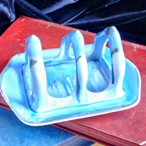 May include: A blue ceramic toast rack with three arched slots, resting on a red book. The rack has a rectangular base and is painted with shades of blue and white. The background features a dark blue velvet fabric.