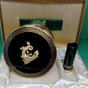May include: A vintage compact mirror and lipstick set in a green box. The compact has a black background with a gold design of a mythical creature. The lipstick is black with gold accents.