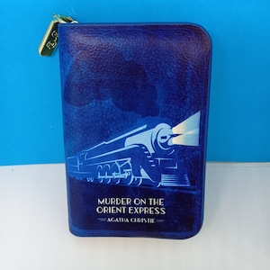 以下が含まれることがあります： アガサ・クリスティの「オリエント急行殺人事件」のタイトルが記された青いブック型ケース。白いスタイリッシュな列車のイラストが表紙に描かれています。金色の留め金でケースが固定されています。