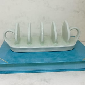 May include: White ceramic bread rack with four slots and a handle on each end. The rack is sitting on a blue book.