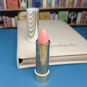 Op de afbeelding: Een roze lippenstift in een zilveren huls, met een wit en goudkleurig patroon lippenstiftkoker op de achtergrond. De lippenstift is open en onthult de roze kleur. Een crèmekleurig notitieboek ligt achter de lippenstift.