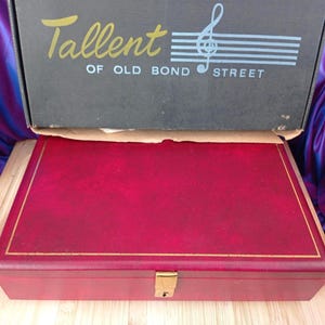 Puede incluir: Una caja rectangular roja con un borde dorado y un cierre dorado. La caja está debajo de una caja negra con el texto "Tallent OF OLD BOND STREET" y un símbolo de nota musical.