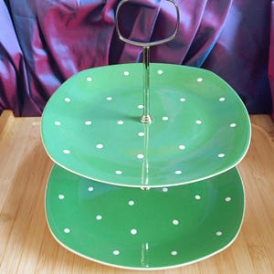 Può includere: Un'alzata a due piani composta da due piatti quadrati verdi con angoli arrotondati e pois bianchi. I piatti sono collegati da un'asta centrale in metallo argentato con una maniglia ad anello coordinata sulla parte superiore.