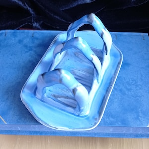 May include: A light blue ceramic butter dish with a handle. The dish is rectangular with rounded corners and a glossy finish. The handle is a series of curved arches. The dish is on a blue surface.