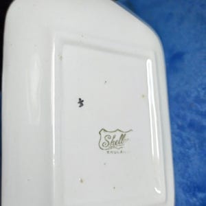 May include: A white, rectangular dish with rounded edges. The dish has a small black mark and a logo that reads "Shell" with "ENGLAND" underneath. The dish is likely made of ceramic or porcelain.