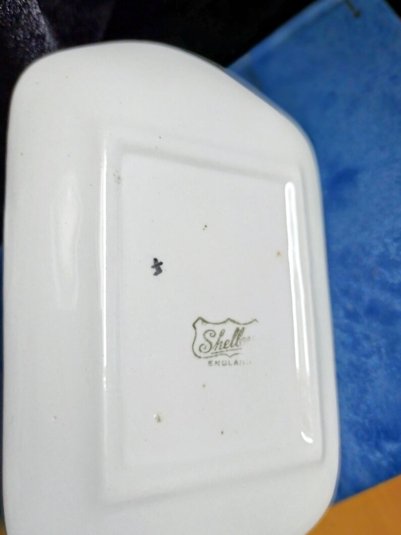 May include: A white, rectangular dish with rounded edges. The dish has a small black mark and a logo that reads "Shell" with "ENGLAND" underneath. The dish is likely made of ceramic or porcelain.