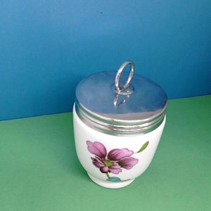 May include: A white porcelain egg coddler with a metallic silver lid featuring a circular ring handle. The body of the container is decorated with a single purple flower illustration with green leaves and a small blue accent at the base of the stem.