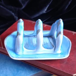 May include: A ceramic blue and white spoon rest with three upright supports. The spoon rest has a rectangular base and is painted with a marbled effect. The background includes a red book and a dark blue velvet cloth.