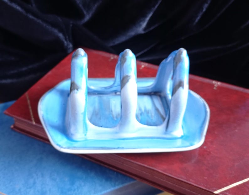 May include: A ceramic blue and white spoon rest with three upright supports. The spoon rest has a rectangular base and is painted with a marbled effect. The background includes a red book and a dark blue velvet cloth.