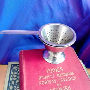 May include: A stainless steel tea strainer with a handle. The strainer has a perforated bowl and a small footed base. The handle is flat with a hole at the end. The strainer is resting on a red book with the text "COOK'S TOURISTS' HANDBOOK NORWAY, SWEDEN AND DENMARK."