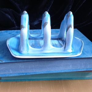 May include: A ceramic toast rack with a blue and white marbled design. The rack has three vertical supports and a rectangular base. The rack is sitting on top of two blue books.