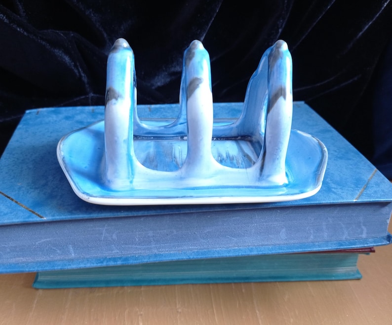 May include: A ceramic toast rack with a blue and white marbled design. The rack has three vertical supports and a rectangular base. The rack is sitting on top of two blue books.