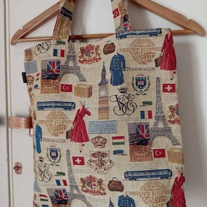 May include: A beige tote bag featuring a woven tapestry pattern of travel-themed imagery, including the Eiffel Tower, Big Ben, Orient Express trains, national flags, vintage suitcases, crests, and figures in red coats and blue overcoats.