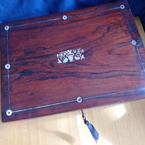Puede incluir: Una caja rectangular de madera de color marrón oscuro con un patrón de veta pronunciado. La tapa está decorada con una incrustación floral blanca central y un borde metálico fino acentuado por pequeñas incrustaciones blancas circulares.