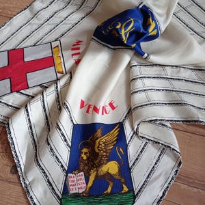 Könnte beinhalten: Elfenbeinfarbener Seidenschal mit schwarzen Streifen und Städtenamen in Rot. Mit einem blau-goldenen Emblem, einer rot-weißen Flagge und einem blau-gelben Design. Der Schal ist gefaltet und zeigt die verschiedenen Muster.