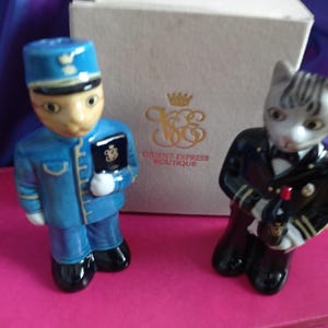 May include: Two ceramic figurines: a cat dressed as a train conductor and a cat dressed as a bellhop. The bellhop cat wears a blue uniform and holds a book. The conductor cat wears a black uniform and holds a bottle. The box behind them says "ORIENT EXPRESS BOUTIQUE".