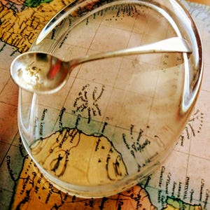 Puede incluir: Un pisapapeles de cristal transparente con forma ovalada que presenta un mapa antiguo detallado del Océano Atlántico y las costas circundantes, coronado con una pequeña cuchara de metal plateado pulido que descansa sobre la superficie.