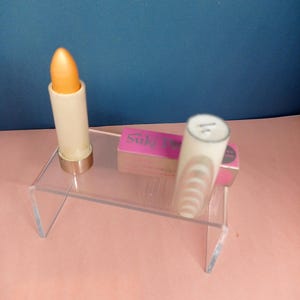 以下が含まれることがあります： 透明なアクリルディスプレイに2本の口紅。1本はオレンジゴールド色で、もう1本はデザインのある白いチューブです。 「Suki」という文字が入ったピンク色の箱もディスプレイにあります。背景は青とピンクです。