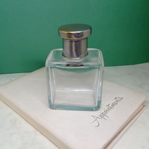 May include: A clear, square glass bottle with a silver-colored cap. The bottle sits on a cream-colored book with the word "Appointments" written on it. The background is a solid green color.
