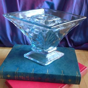 Puede incluir: Un cuenco de vidrio transparente azul claro con base cuadrada y diseño geométrico acampanado. El cuenco está sobre un libro azul oscuro con detalles dorados, apoyado en un libro rojo. El fondo presenta una tela morada drapeada.