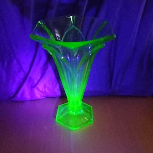 May include: A clear glass vase with a vibrant green glow. The vase features a flared, petal-shaped rim and a hexagonal base. The glass has a ribbed design, and the vase is lit with a green light.