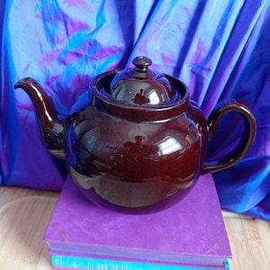 May include: A glossy, dark brown ceramic teapot with a lid, spout, and handle. The teapot is sitting on a stack of purple books. The background is a blue and purple fabric.