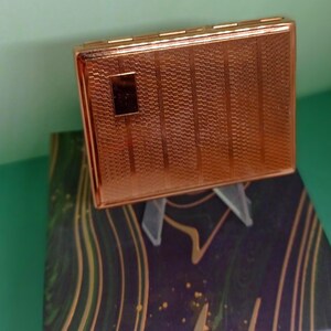 May include: A rectangular, rose-gold colored cigarette case with a textured, patterned surface. The case has a small, square accent on the front. It rests on a decorative box with a purple and gold design.