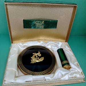 May include: A green and gold compact mirror with a dragon design and a matching lipstick tube. The compact is in a velvet-lined box with the words "Vogue Gemities Made in Ireland" on the inside.