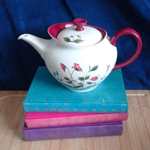 May include: A white ceramic teapot featuring a magenta handle, a matching magenta lid knob, and a floral pattern of pink flowers with green leaves. The teapot has a curved spout and sits atop a stack of three books in teal, pink, and purple.