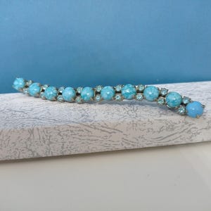 Puede incluir: Un brazalete vintage curvado con piedras turquesas alternas, grandes y pequeñas. Las piedras más grandes tienen un aspecto veteado, y las más pequeñas son transparentes. El brazalete está engastado en metal plateado y descansa sobre una superficie blanca texturizada.