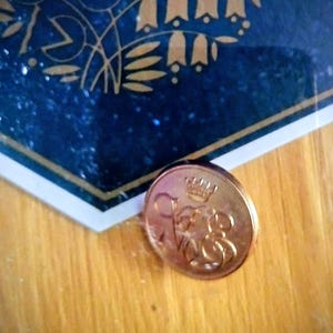 Puede incluir: Una moneda de color cobre con una corona e iniciales ornamentadas. La moneda está sobre una superficie de madera, cerca de un diseño azul y dorado.