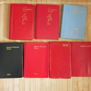 May include: A collection of seven vintage Letts diaries from the 1970s, featuring various red, blue, and black covers. The books are labeled with years ranging from 1970 to 1977, with some covers displaying the text Letts Schoolgirls Diary.