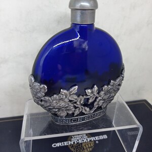 May include: A cobalt blue glass bottle with a rounded shape and a silver-toned metal cap. The base of the bottle is encased in an ornate silver-toned metal floral filigree overlay featuring the embossed text VENICE SIMPLON ORIENT-EXPRESS.