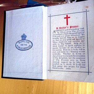 May include: An open book with white pages featuring a red cross and the text A Sailor's Prayer. The left page displays a blue oval stamp reading Supplied for the Public Service with a crown icon. The right page contains black printed text.