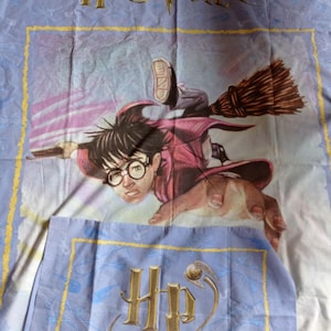 May include: A light blue bed sheet with a graphic of Harry Potter on a broomstick, and the words "Harry Potter" in gold. The bottom section shows the "HP" logo in gold, a design from the popular book series.