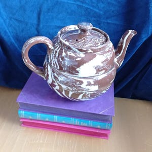 Può includere: Una teiera in ceramica marmorizzata marrone e bianca con coperchio e manico poggia su una pila di tre libri. I libri sono viola, turchese e rosa. La teiera ha un beccuccio e un piccolo pomello sul coperchio.