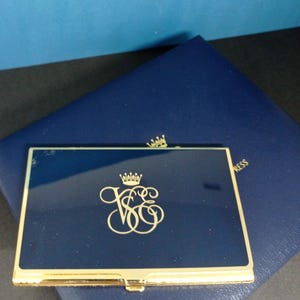 May include: A navy blue business card holder with a gold frame and accents. The holder features a gold crown and monogram design. It rests on a navy blue leather-like surface.