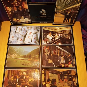 May include: A collection of vintage postcards featuring scenes from the Venice Simplon-Orient-Express. The postcards depict train interiors, landscapes, and people. The set includes a black box with gold lettering and a logo.