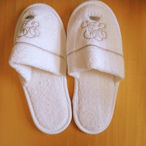 May include: A pair of white terry cloth slippers with embroidered detailing. Each slipper features a crown and monogram design in a light green and tan color. The slippers are placed on a light brown wooden surface.