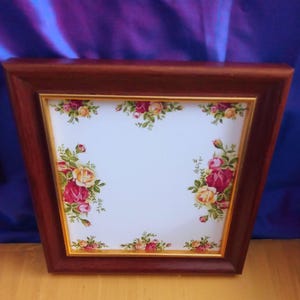 Puede incluir: Un marco de madera cuadrado con un acabado marrón oscuro que rodea un azulejo blanco. El azulejo presenta un patrón floral con racimos de rosas rojas, rosas y amarillas con hojas verdes dispuestas a lo largo de los cuatro bordes de la superficie.