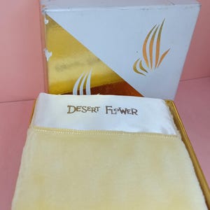May include: A vintage "Desert Flower" powder box. The box is white with gold accents and a stylized floral design. Inside, a fluffy, yellow powder puff rests on a white cloth, with the brand name printed on it. The box is rectangular.