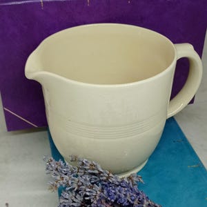 May include: A white ceramic jug with a handle and a slightly flared lip. The jug has a smooth, cream-coloured glaze and a subtle embossed design. The jug is sitting on a blue surface with a bunch of dried lavender flowers.