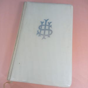 May include: A cream-coloured book with a light blue monogram on the cover. The book has a ribbon bookmark and is slightly open, revealing the pages. The cover is glossy and the edges are rounded.