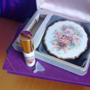 以下が含まれることがあります： 花束のプリントとゴールドの縁取りが施された白いコンパクトミラー、ゴールドのキャップと花柄が特徴の小さな白い香水瓶、そして透明なプラスチック製の小さな漏斗が、グレーのベルベットが敷かれた収納ケースの中に収められています。